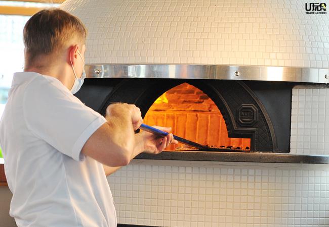 厨师正将披萨放入意大利进口的Alfa Forni燃木和煤气烤炉进行烘制。它不仅是意大利卓越工艺的标志，也是马来西亚少见的披萨专业烤炉。